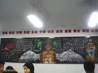 这是一张拍摄于教室的照片，展示了一块清明主题的手绘黑板报。黑板报中心绘制了带有天使光环和白色翅膀的科比·布莱恩特形象，搭配绿色的“清明”大字，将清明节的追思氛围与对科比的缅怀结合；左侧绘制红色直升机图案，配有缅怀科比因直升机事故离世的文字内容，右侧则绘制冰红茶饮料图案，同样配有清明时节追思科比的文字。教室墙面上还印有红色的社会主义核心价值观标语，画面左下角有拍摄时间水印“2024/04/02 21:20:39”，下方能看到部分学生的身影，整体是校园里以创意方式进行清明纪念的场景。