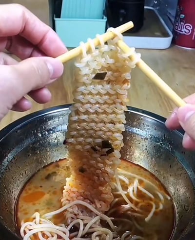 这张静态图片呈现了一个趣味视觉错位场景，画面中两只真人的手分别持一双筷子，共同夹起一撮泡好的方便面。由于方便面面饼本身的网格状纹理，加上筷子的摆放角度，使得被夹起的部分看起来如同被编织成了带有规律针织纹路的织物，视觉效果新奇有趣。下方的金属碗中盛放着剩余的泡开面条和混有调料的褐色汤汁，汤汁里还能看到少许调料碎末。背景是木质桌面，上面摆放着浅蓝色收纳盒、黑色容器和红色包装物品等杂物，整体是日常居家的用餐场景，利用视觉错觉为普通的吃方便面场景增添了创意趣味。