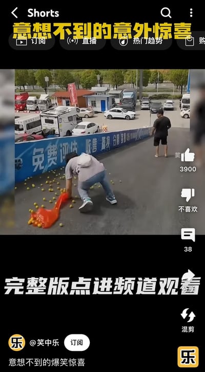 这是一段竖屏短视频的截图，画面呈现了户外停车场旁的路面场景：一名穿浅灰色上衣的人正蹲在地上，试图捡起撒落一地的黄色果子，装果子的橙色袋子还放在身旁；不远处站着一名黑衣男子。背景里的停车场停着房车、轿车、货车等多种车辆，蓝色围栏上印有“免费评估 收售 置换 分期 寄售”的广告字样。视频顶部配有文字“意想不到的意外惊喜”，底部标注“完整版点进频道观看”，账号简介为“意想不到的爆笑惊喜”，整体是主打搞笑效果的生活意外类短视频，暗示后续会有反转的“惊喜”情节。