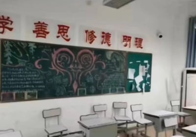 这是一张教室内部场景的照片。教室墙壁为白色，上方悬挂着红色的中文标语，内容从左到右依次为“学”“善思”“修德”“明理”。正前方是一块大型黑板，上面有彩色粉笔绘制的图案，包括一个显眼的粉色心形花纹，周围还有一些文字和装饰性图案，黑板右侧部分为绿色背景，贴有白色的纸张拼贴。教室里摆放着多排白色的课桌椅，右侧有一扇带有蓝色窗帘的窗户，墙上安装有一个监控摄像头。整体环境整洁，呈现出典型的校园学习氛围。
