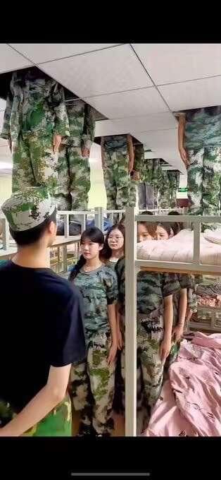 这张图呈现了军训宿舍里的趣味视觉错位场景：下铺区域站着几位身穿军训服的女生，而上铺床板边缘悬挂着几条迷彩长裤，恰好与下铺女生的上半身在视角上重合，形成了“女生没有头部，只剩身体”的荒诞搞笑效果，利用视角巧合制造出充满趣味的视觉错觉。