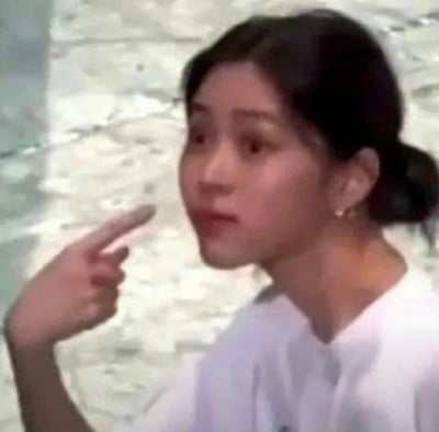 这是一张静态真人表情素材图，画面中的女性将头发盘在脑后，身着白色上衣，背景是浅色的石质墙面。她眼睛瞪大，面部呈现出惊讶的神情，同时伸出一只手，用食指和中指指向前方，整体姿态和表情传递出震惊、不敢相信甚至带有指责意味的情绪，非常适合在社交聊天中用来表达对某件事的意外、吐槽或是不满的态度。