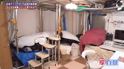 图片展示了一个杂乱的房间场景，可能是日本某家店铺的内部。房间内有金属框架结构，一个覆盖着黑白图案布料的大型充气物体（疑似床或沙发），旁边有几个小型折叠凳，地面铺着拼接地垫。右侧有一张书桌，上面放着电脑显示器和其他杂物，周围堆放着布料、袋子等物品。顶部悬挂着灯具和绿色软管，整体环境显得拥挤且充满生活气息。左上角有日文文字：“日本でここだけ!? お店の珍百景 アナタは体験したいと思うか？”（意为“只有在日本这里吗!? 店铺的罕见景象 你想体验一下吗？”），右下角有“珍百景”水印。