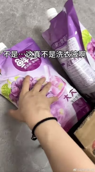 图片中展示了两袋紫色包装的饮料，包装袋上印有饱满的葡萄图案和“果味爽 葡萄味”等字样，一只戴着黑色手环的手正轻轻按在其中一袋的正面。图片上方配有醒目的文字“不是…这真不是洗衣液啊”，幽默地调侃该饮料包装因颜色和形状与洗衣液过于相似而容易引发的误会。场景真实接地气，通过日常物品的视觉混淆制造出轻松搞笑的效果。