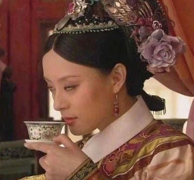图片展示了电视剧《甄嬛传》中的经典场景：主角甄嬛身着华丽的清代宫廷服饰，头戴镶嵌珠宝与紫色花朵的精致凤冠，正手持一个绘有花纹的小巧茶杯，低头轻啜茶水。背景为红色帷帐，整体呈现出宫廷环境的庄重与典雅氛围。