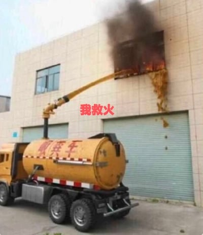 这张图实拍了一处滑稽的乌龙救火现场，浅米色外墙的工业厂房二层的一扇窗户正在起火燃烧，窗口冒出大量黑色浓烟，一辆黄色吸粪车停在厂房门前，车上的机械长臂将出料口对准起火窗户，正往起火点喷出黄褐色的粪便，大量粪便顺着墙面流到下方的卷帘门上，图片中间位置添加了红色文字“我救火”，吸粪车罐体上也标注了红色的“吸粪车”字样，整体场景意外又搞笑。
