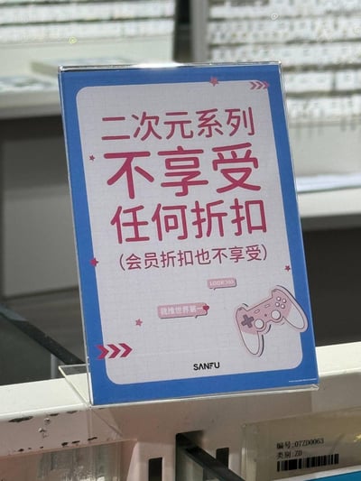 这是一张 retail 店内的提示牌照片，展示了一个蓝色边框的亚克力标牌。标牌中央有醒目的粉色文字'二次元系列 不享受任何折扣'，下方还有补充说明'(会员折扣也不享受)'。标牌设计包含卡通风格的游戏手柄图案和装饰性星星、箭头元素，底部有'SANFU'品牌标识。背景显示了 retail 店的货架环境，表明这是在实体店内使用的商品政策提示牌。