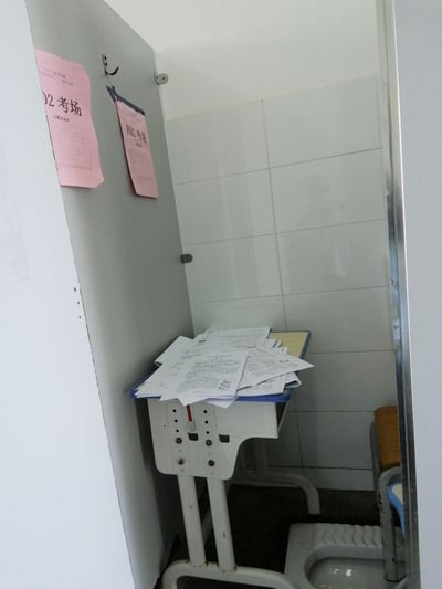 这张图片展示了一个令人意外的场景：在一个带有蹲便器的卫生间空间里，靠墙放置着一张堆满试卷的学生课桌，旁边的门上贴着“02考场”和“F02考场”的粉色标识，墙面铺着白色瓷砖。这种将考场相关的试卷和考场标识出现在卫生间的场景，显得十分反常，给人一种荒诞、离谱的感觉，似乎是考场布置中的特殊情况或者是让人哭笑不得的安排。