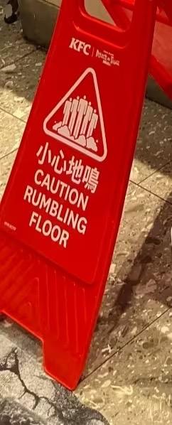 这是一张经过P图改编的KFC门店警示牌照片，原本的“小心地滑”被修改为“小心地鳴”，英文对应改为“CAUTION RUMBLING FLOOR”，放置在带有水渍的地面旁。

使用了谐音梗：将“小心地滑”的“滑”替换为“鳴（鸣）”，搭配英文的“RUMBLING FLOOR”，制造出趣味效果，调侃地板仿佛在震动发出声响，让普通的警示牌变得搞笑有趣。