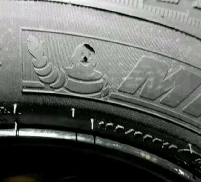 这张图片展示了轮胎胎面的特写镜头，胎面上有一个有趣的图案设计，看起来像一个卡通形象。图案由轮胎的花纹构成，包括类似头发的螺旋状设计和一个圆形元素，而圆形元素中间恰好有一个小孔，形成了类似眼睛的效果。胎面上还能看到部分品牌标识，显示出字母"M"。