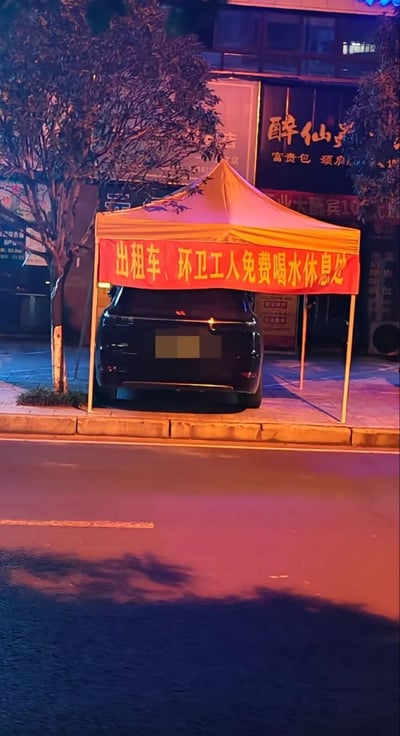 这是一张夜晚拍摄的街头实景照片，画面里人行道上搭建了一座橙黄色临时帐篷，帐篷正面悬挂红色横幅，标注此处为出租车司机、环卫工人免费喝水休息的公益服务点。但帐篷下方的全部空间被一辆黑色私家车违规停放占据，完全挤占了本应提供给户外劳动者的休憩区域，场景极具讽刺性，反映了公共公益资源被私人占用的不文明现象。背景还可见周边的商铺招牌和行道树，整体被夜晚街灯映照出暖橙与暗蓝交织的光线。