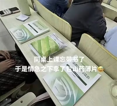 课堂场景的课桌特写，桌面上摆放着两本绿色封面的课本，旁边显眼地放着一包绿色包装的山药薄片。背景中能看到其他同学坐在座位上，似乎正在上课。图片配文“同桌上课忘带书了 于是情急之下拿了包山药薄片😂”，描绘了同学因忘带课本而用零食包装应急的搞笑场景，充满校园生活的趣味和幽默感。