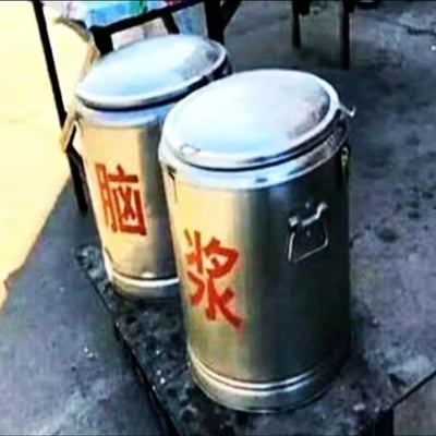 这是一张原始拍摄的静态图，画面展示了放置在室外水泥台面上的两个不锈钢保温桶，这类保温桶通常被用来盛装豆浆、粥品等早餐食物。但桶身分别用红色字体标注了“脑”和“浆”，将原本常见的“豆浆”类食物名称拆分重组，制造出“脑浆”这个带有惊悚感的词汇，形成了强烈的反差效果，属于生活中的趣味恶搞创作，通过打破常规认知来制造搞笑的视觉趣味。