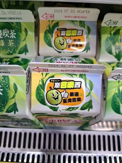 这是在超市冷藏货架上拍摄的斯芭樂西台湾芭乐风味盒装饮料，包装上印有竖起大拇指的拟人化芭乐卡通形象，配文“すばらしい 斯芭樂西 嚴選 臺灣芭樂”，包装顶部标注了有效日期，如2024.06.27、2024.07.01，旁边还摆放着纯喫茶绿茶类饮料，整体呈现出便利店冷藏柜的商品陈列场景，没有双关或谐音梗内容。