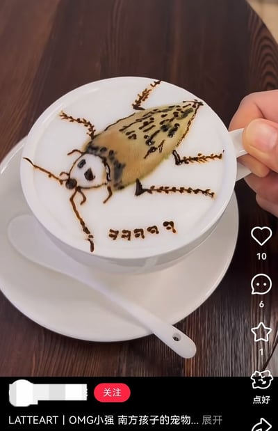 图片展示了一杯拿铁咖啡，其奶泡上用咖啡绘制了一只卡通化的蟑螂形象。蟑螂有着长长的触角，形似双马尾，这是对"广式双马尾"网络热梗的创意改编。蟑螂的面部被绘制得较为可爱，带有圆圆的眼睛和微笑的表情。杯子被一只手握着，放在木质桌面上，旁边有一个白色的勺子。图片底部有文字"LATTEART | OMG小强 南方孩子的宠物..."，暗示了这是南方地区对蟑螂的一种幽默调侃。