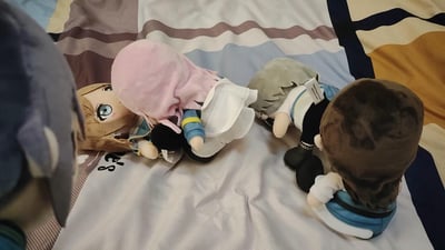 图片展示了多个动漫风格的 plush 玩具被摆放在铺有浅色床单和条纹毯子的床上。这些 plush 拥有不同的头发颜色，包括粉色、灰色、棕色和金色，姿态各异，有的侧卧，有的平躺，营造出温馨可爱的氛围，像是动漫爱好者的个人收藏展示。