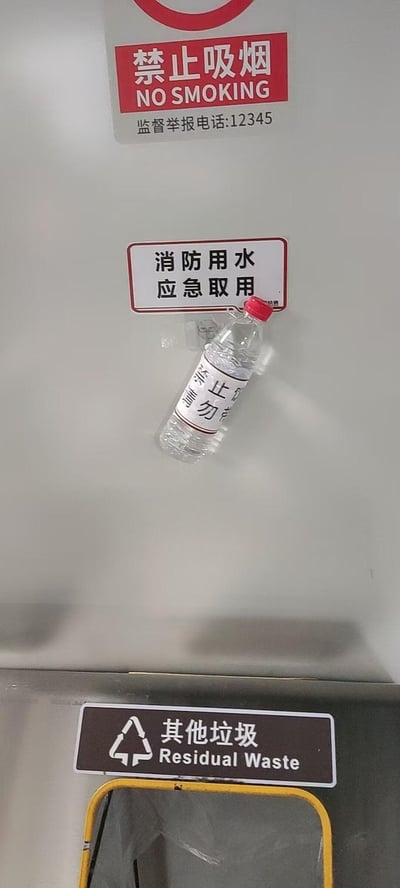 这是一张竖拍的真实公共场景照片，画面从上到下依次呈现：
1. 顶部红底白字的「禁止吸烟」标识，标注英文“NO SMOKING”及监督举报电话12345；
2. 中间白底黑字的「消防用水 应急取用」标识，其下方的出水口被一个贴有「禁止饮用 请勿」字样标签的矿泉水瓶套住，形成了“把应急消防水装进矿泉水瓶”的滑稽反差感；
3. 底部是「其他垃圾 Residual Waste」的垃圾分类标识。

画面没有双关、谐音梗，只是记录了现实里有点乌龙的有趣瞬间。