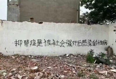 一张拍摄于城市废墟中的照片，画面中央是一面斑驳的白色墙壁，上面用黑色喷漆写着极具争议性的文字："抑郁症是被社会强奸后感染的性病"。墙壁下方是散落的砖石瓦砾，背景中可见一栋灰色的残破建筑和部分绿树。整个场景呈现出一种破败、压抑的氛围，与涂鸦内容所表达的社会批判形成呼应。