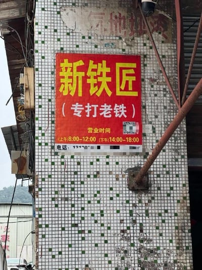 这是一张线下街边小店的竖版实拍图，画面核心是贴在带有绿白小方格瓷砖的老旧墙柱上的红色店铺招牌。招牌以黄色大字写着“新铁匠”，下方用白色字体标注“（专打老铁）”，还列出了营业时间“(上午)8:00-12:00 (下午)14:00-18:00”以及部分被打码的联系电话。这里的“专打老铁”是巧妙的双关：一方面契合铁匠铺打铁的本职工作，另一方面“老铁”是网络语境中对朋友的亲昵称呼，用网络热词玩梗让普通的店铺招牌充满趣味和网感。墙柱上方残留着旧招牌的模糊痕迹，周围可见杂乱的管线和外部的简易建筑，整体呈现出接地气的城镇街边小店风貌。