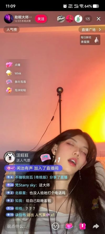 该图片为某短视频平台的直播画面截图，主播“助眠大师”正躺卧着，双眼闭合，佩戴耳机，背景是橙红色渐变灯光，前方有麦克风支架。直播界面左侧显示“点播”“Wink”等互动图标，右侧有“人气榜”“直播广场”等入口，底部滚动着用户评论，如“给自己助眠着呢”“这大师”等，画面整体呈现放松的直播氛围。