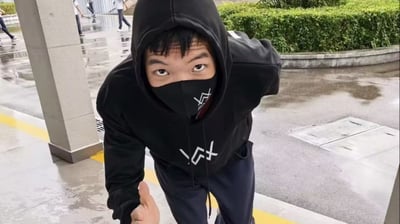 这张实拍图拍摄于雨后的校园室外公共区域，地面留有明显积水，远处可见多名穿着校服的学生走动。画面主体是一名年轻男性，他穿着印有类似欧美DJ Alan Walker标志性标识的黑色连帽卫衣，戴上卫衣帽子遮挡住大半头部，同时佩戴同风格的黑色印花口罩，仅露出双眼看向镜头，身体微微前倾，对着镜头伸出手比出点赞的手势，刻意模仿Alan Walker的全黑造型，呈现出中二耍帅的状态，是网络热梗「嘉豪」的代表性素材。