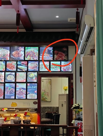 这是一张中式餐馆内部的实拍照片，墙面上方排列着多块发光的菜品展示灯箱，展示了多种不同的餐品。右上角的一块灯箱菜品图被红色圆圈特意标注，该图内容为红亮的卤制大肘子，因色泽、造型和视觉角度的原因，乍看会被误认成红色的处于狂笑状态的奶龙形象。画面下方可见餐馆的用餐桌椅、柜台标识，还有通往内部区域的门洞，整体是常见的中式家常菜馆的店内环境。