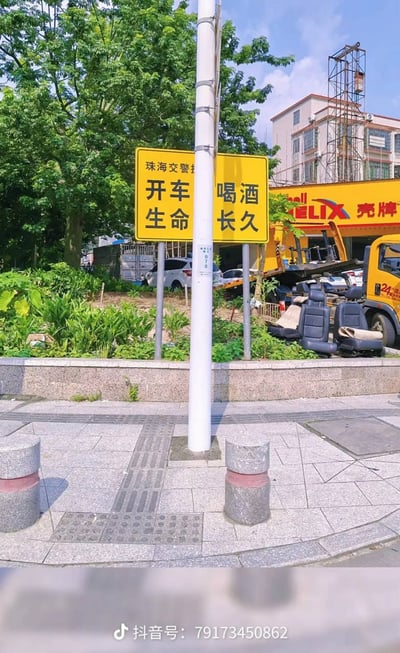 这是一张实拍的街景照片，画面中央的白色电线杆恰好挡在两块相邻的黄色珠海交警警示路牌中间，原本宣传“开车不喝酒，生命更长久”的安全标语被遮挡后，视觉上呈现出“开车 生命”“喝酒 长久”的拆分效果，很容易被误读为含义完全相反的搞笑内容。画面背景可见路边的绿植、壳牌门店、黄色工程车辆、停放的私家车以及在建的居民楼，前景是人行道的石材路面和隔离石墩，图片底部带有抖音发布者的账号水印。