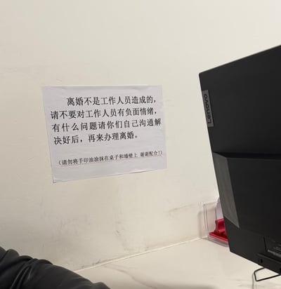 这张图片实拍了民政部门离婚办理区域的墙面场景，墙上贴着一张白底黑字的提示标语，全文为：
> 离婚不是工作人员造成的，请不要对工作人员有负面情绪，有什么问题请你们自己沟通解决好后，再来办理离婚。（请勿将手印油涂抹在桌子和墙壁上 谢谢配合！）
画面右侧露出一台联想（Lenovo）显示器的部分机身。标语内容直白地透露出离婚窗口工作人员常被迁怒的无奈处境，括号里的补充说明更是暗示了此前出现过的离谱行为，真实又带着一丝戏谑感。