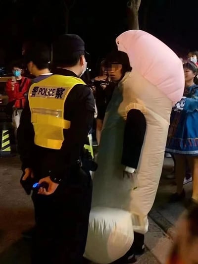夜晚的街道上，一名穿着黄色反光背心、印有“警察POLICE”字样的警察正在与一名身着巨大粉色和白色充气服装的人员交流。周围有几位戴着口罩的路人在围观，有人正在用手机拍照。场景看起来像是在某个公共活动或节日期间，警察正在对特殊装扮的人员进行例行询问或检查。