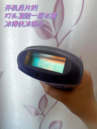一张真人手持深色手持设备的原始拍摄图，设备灯头处可见因制冷产生的彩虹色水雾凝结现象，背景为带有紫色花卉图案的白色壁纸。图片左上角有粉色渐变文字标注：'开机后片刻 灯头凝结一层水雾 冰得快冰得久'，直观展示设备制冷性能。整体构图清晰，通过实物效果与文字结合进行产品功能宣传。