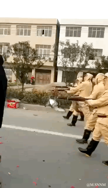 这是一段动态GIF内容，画面中一群穿着类似日军军装的表演者排成队列，手持道具枪做出瞄准射击的动作，地面散落着红色的鞭炮碎屑，他们通过鞭炮爆炸的效果来模拟开枪的声响与视觉效果，背景是城镇街道和居民楼，整体是充满趣味的民间自娱自乐表演，氛围轻松搞笑，仅为趣味模仿无恶意。