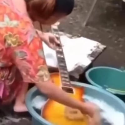 图片中一名身穿红色花纹连衣裙的女子正将一把黄色电吉他完全浸入蓝色水盆中清洗，盆内有白色泡沫。她赤脚站在灰色水泥地面上，身旁还放着另一个绿色水盆和一块白色抹布。这一场景因电吉他属于精密电子乐器，通常需专业清洁而非水洗而显得极具反差感和迷惑性。