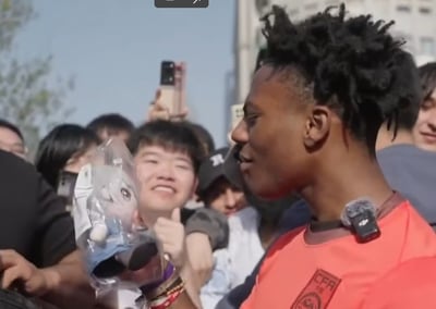 图中是知名美国游戏主播IShowSpeed（伊曼纽尔·安德森）在中国参与线下活动时与粉丝互动的场景。他身着带有CFR标识的橙色上衣，衣领佩戴着领夹麦克风，侧头面向身边的粉丝，面带笑意。周围簇拥着多位热情的粉丝，其中一位粉丝笑着举着包装好的玩偶，还竖起大拇指，现场氛围十分热烈，尽显粉丝对这位海外主播的喜爱与欢迎。