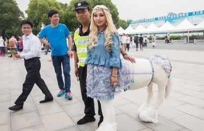 在户外的广场活动现场，一位装扮成半人马的coser站在画面中央，她有着金色长卷发，头戴花环头饰，上身穿着蓝色荷叶边短上衣，通过道具与站位营造出半人马的视觉错位效果——白色的马身道具连接在她身体后方，搭配白色毛绒裤脚，整体造型奇幻有趣。一旁穿着带有“SECUR”标识反光背心的安保工作人员，将手搭在马身道具上，似乎是好奇地与coser合影。背景中还有路过的行人，有的面带笑意看向镜头方向，远处是白色帐篷和写有“大力发展 抢占文化产业发展制高点”的蓝色横幅，现场氛围轻松热闹。这个半人马造型是通过道具和巧妙站位形成的视觉错觉，并非真实的身体结构。