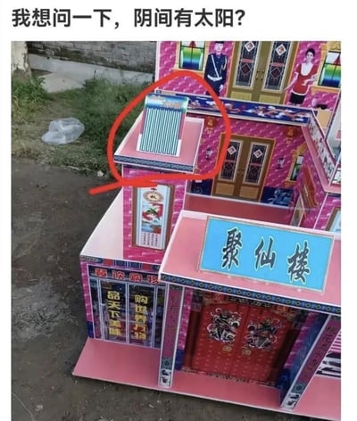 图片展示了一个户外场景中的纸扎建筑模型，整体为粉色调，外观类似传统中式别墅，建筑上方有“聚仙楼”的蓝色牌匾，侧面还可见“品天下美味”“购物广场”等字样。模型屋顶上安装了一个太阳能热水器，被红色圆圈重点标注。图片顶部配有文字“我想问一下，阴间有太阳？”，通过传统殡葬祭品（纸扎屋）与现代生活用品（太阳能热水器）的组合，形成幽默反差，引发对“阴间是否需要太阳能”的调侃式疑问。