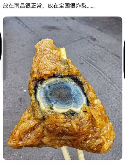 这是一张顶部加了配文的美食实拍图，顶部配文为“放在南昌很正常，放在全国很炸裂......”，画面主体是一颗被切开的三角咸粽，酱色的调味糯米内部包裹着一整颗完整的皮蛋，粽子被竹签串起，背景为灰色水泥地面。这种将整颗皮蛋作为粽子内馅的做法是南昌当地的特色饮食，对于国内其他地区的人群来说较为少见，会感到新奇甚至意外。