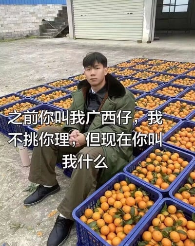 图片中一名穿着绿色外套的年轻男子坐在满地蓝色塑料筐的橘子堆中，背景是灰色水泥地和卷帘门仓库。图片上方覆盖白色文字：“之前你叫我广西仔，我不挑你理，但现在你叫我什么”。通过幽默的文字对比，暗示男子因拥有大量橘子而身份或境遇发生变化，引发对新称呼的调侃，整体氛围轻松诙谐。