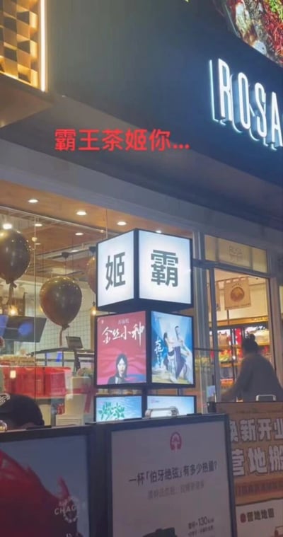 夜晚的商业街区，一家疑似'霸王茶姬'的茶饮门店外，招牌灯箱意外将'姬'与'霸'二字上下排列，形成醒目的'姬霸'组合。门店上方红色电子屏滚动文字'霸王茶姬你...'，搭配下方错位的招牌文字，引发路人会心一笑。店内可见装饰气球与电子菜单屏，隔壁为'ROSA'品牌店铺，整体呈现热闹的都市商业夜景。