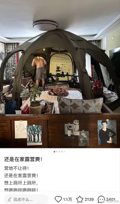 这是一张社交媒体截图，展示了一种另类的“居家露营”生活方式：在自家客厅里搭建起大型露营帐篷，帐篷内布置了桌椅、绿植、咖啡器具等露营相关物品，一名赤裸上半身的男子在帐篷内进行整理。下方配文调侃因户外营地无法停留，转而选择在家“露营”，突出这种方式的便捷性——可以随时上厕所、喝咖啡，幽默地对比了户外露营的不便与居家“露营”的舒适惬意，传递出一种无奈又乐观的生活态度。