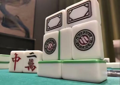 图片展示了一副麻将牌的创意堆叠场景。前景中，“中”和“二万”两张牌平放在绿色桌布上，后方则堆叠着数张麻将牌，其中可见带有圆形图案的“三筒”牌，整体构图清晰，展现了麻将牌的经典样式与质感。背景隐约可见一幅画作，增添了场景的生活气息。