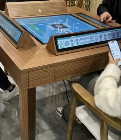 这是一张实拍的新式智能麻将桌照片，桌子整体为木质设计，桌面中央是一块大型触控显示屏，展示麻将对局的公共牌面，桌子四边各对应一位玩家的位置，分别安装了倾斜的触控显示屏，用来展示每位玩家的私有手牌，完全替代了传统的实体麻将牌，实现全电子化的麻将娱乐。画面中可见三位玩家的部分身体：右上角有玩家正用手指点击侧边屏幕操作，右侧有穿着白色毛绒外套的玩家手持手机，左下角露出了另一位玩家的鞋子，整体场景是在室内体验这款智能麻将设备。