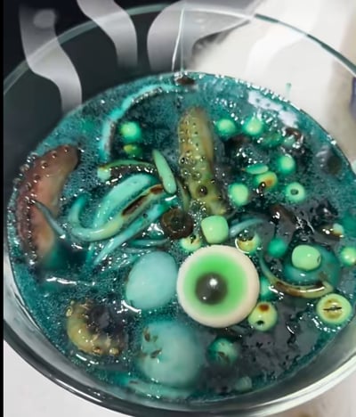 这是一款为万圣节打造的创意黑暗料理，玻璃碗中盛着蓝绿色带泡沫的诡异汤汁，里面漂浮着仿真章鱼触手、造型逼真的眼球状糖果，还有其他仿海鲜食材，整体视觉效果充满惊悚猎奇感，完美贴合万圣节的搞怪氛围，用来烘托节日的诡异气氛。