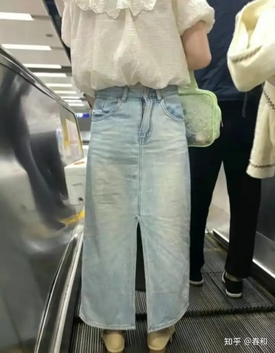 这张图片拍摄于公共场所的电扶梯上，一位女生穿着白色泡泡袖上衣和淡蓝色牛仔长裙，从后面看因为走路姿势和裙子的褶皱、光影效果，造成了视觉错觉，好像牛仔裙变成了“牛仔裤”，仿佛有两条腿分开。这样神奇的视觉效果令人忍俊不禁，属于网络视觉梗图。

本图没有双关、谐音梗或特殊暗示，单纯依靠视觉误导带来趣味。
