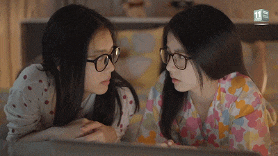 图中是两位戴着黑框眼镜的女生，她们趴在桌前，脑袋紧紧凑在一起，目光专注地盯着面前的屏幕，神情认真又带着好奇，仿佛正在围观什么有意思的内容，完美还原了和好友一起吃瓜、看八卦的日常状态。