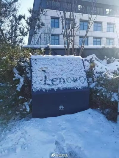 这是一张雪后户外拍摄的趣味照片，画面中心是一块带有华为标志的深色标识牌，标识牌表面的积雪被人用手扫出了“Lenovo（联想）”的品牌字样，形成了品牌错位的搞笑反差效果。背景是标有“Q1”字样的办公楼建筑，周围的低矮植被和地面都覆盖着一层积雪，呈现出冬日雪后的清冷氛围。这个场景通过在华为的专属标识牌上标注竞争对手的品牌名，制造出无厘头的趣味梗，充满了网友的恶搞创意。