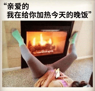 图片上方有文字“亲爱的 我在给你加热今天的晚饭”，下方是一个人双腿朝上、脚穿绿色袜子和灰色裤子，躺在壁炉前的地上，双手拿着手机。壁炉里火焰熊熊燃烧，形成一种幽默的反差——文字说在加热晚饭，实际画面却是人以奇怪姿势躺在燃烧的壁炉前。这张图通过文字与画面的不协调制造笑点，暗示所谓的“加热晚饭”可能只是个借口或玩笑。