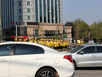 在城市的街头路口，一群身着统一黄色制服、头戴黄色头盔的美团外卖员整齐站立集合，推测是在进行岗前早会或工作安排。前景是两辆停靠的汽车，背景可见招商银行的高层建筑、茶百道门店，周边树木枝叶繁茂，展现出春日城市里外卖从业者的日常工作场景。背景墙面还可见社会主义核心价值观的标语文字。