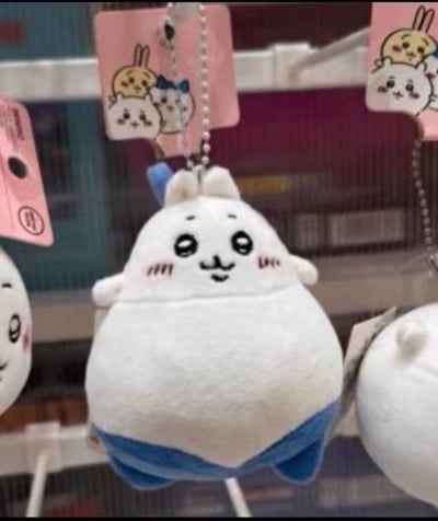 这是日本超人气IP《Chiikawa》（吉伊卡哇）的衍生毛绒挂件，角色为乌萨奇。玩偶整体圆滚滚，身体下半部分为蓝色，头顶带有蓝色小装饰，脸上带着微妙的搞怪笑意，脸颊配有腮红，造型既可爱又滑稽。它被悬挂在展示架上，周围还有同系列的其他玩偶挂件，背景吊牌上印有Chiikawa系列的角色图案。