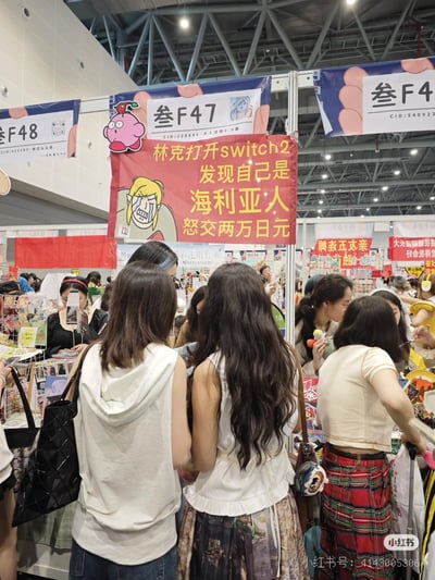 这是一张同人漫展现场的实拍图，画面中叁F47摊位悬挂着红色玩梗横幅，横幅上画着崩溃哭泣的《塞尔达传说》角色林克，配文「林克打开switch2发现自己是海利亚人怒交两万日元」，调侃传闻中的Switch2可能出现的离谱收费设定；摊位上方还贴有星之卡比的卡通贴纸，周围聚集了不少逛展的爱好者，现场氛围热闹，充满同人展会的活力。