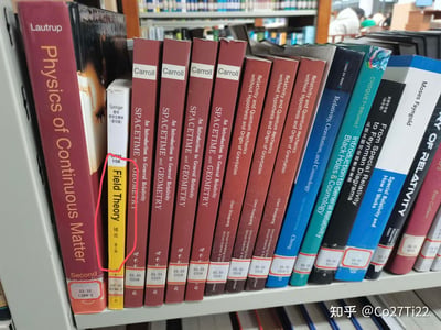 这是一张在图书馆内拍摄的书架实拍图，书架上整齐陈列着多本物理、数学领域的外文专业学术书籍，涵盖连续介质物理、广义相对论、场论等方向的经典教材与专著，其中一本《Field Theory 域论 第2版》被红圈标注出来。背景中可以看到图书馆的其他书架和正在学习的读者，整体氛围安静且充满学术感，图中没有双关、谐音梗或暗示内容。